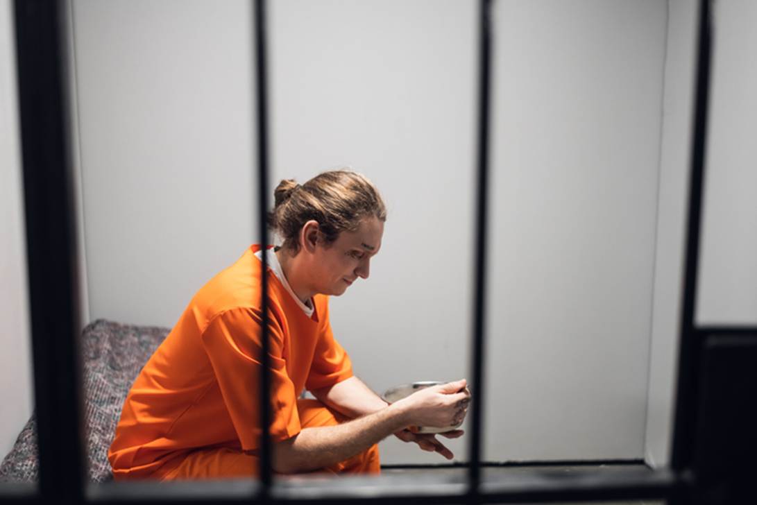 A person sits in a jail cell. The person, wearing an orange jumpsuit, looks down at a bowl in their hands.